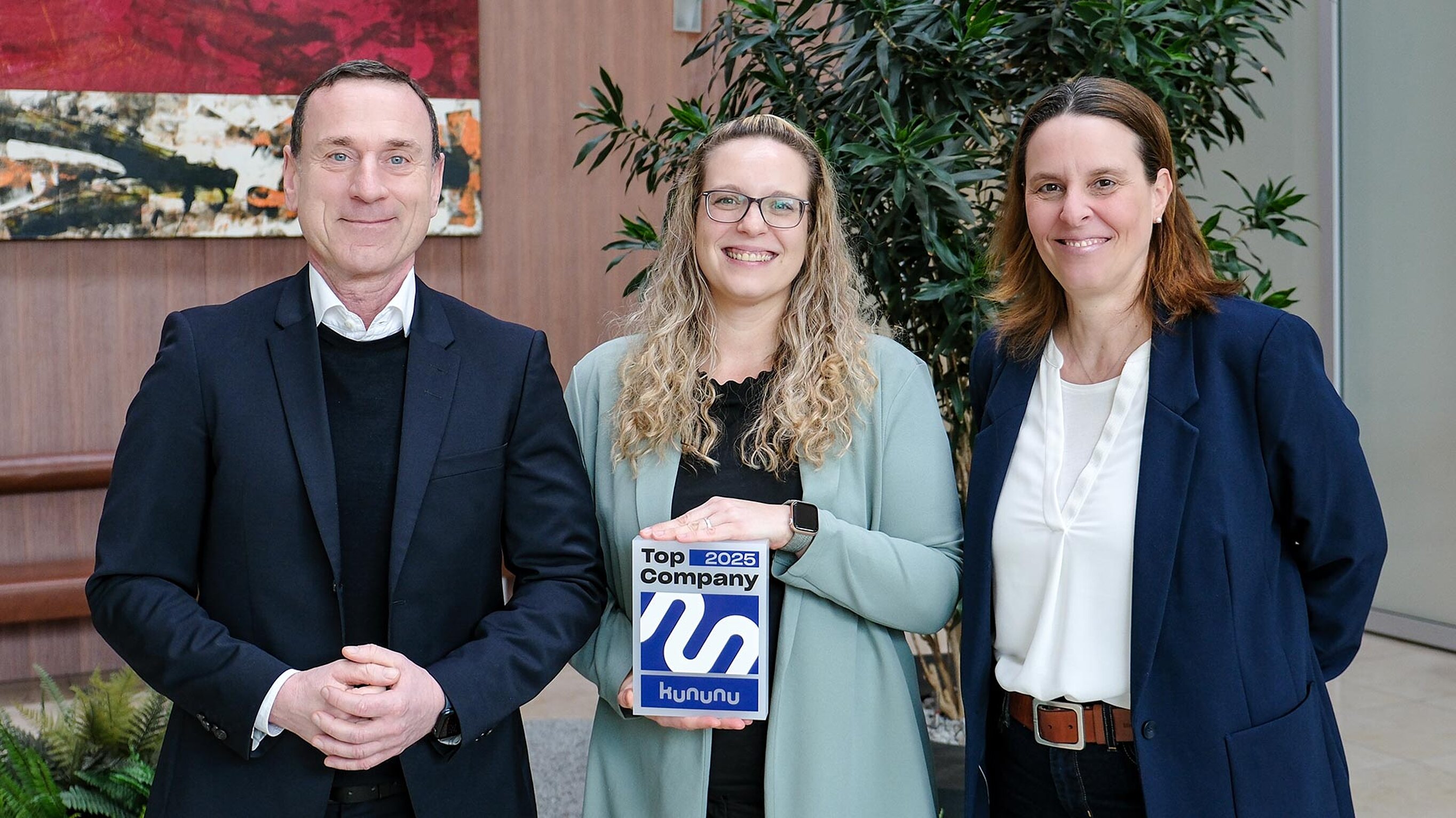 Drei Personen stehen im Foyer nebeneinander. Die Frau in der Mitte hält den Award